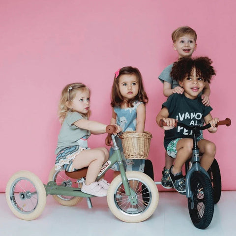 Four young children are posing against a pink background. Two kids are sitting on TryBike - Vintage Green balance bikes, while the other two stand behind them. The children are smiling and wearing casual clothes, with a mix of expressions and hairstyles, enjoying their time learning to ride.