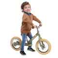 A joyful child with curly hair and a brown sweater is learning to ride a TryBike - Vintage Green balance bike with beige tires. The child is smiling, dressed in blue jeans and black sneakers with white soles, clearly having fun on their TryBike from the brand TryBike.