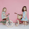 Two young children are standing with their TryBike Steel balance bikes against a pink background. The child on the left wears a grey outfit and holds a green 2-in-1 tricycle balance bike, ideal for learning to ride. The child on the right wears a blue dress with white designs and stands next to her TryBike - Vintage Pink, which has a basket attached.