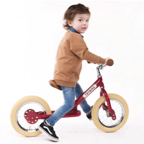 A young child, wearing a brown sweater, blue jeans, and black and white sneakers, is riding a TryBike - Vintage Red 2-in-1 tricycle balance bike with beige tires. The child looks back over their shoulder while balancing on the bike, which has "TRYBIKE" printed on the frame.