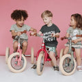 Three young children are sitting on small TryBike Steel bicycles with beige tires against a pink background. They appear to be chatting and having fun. The child on the left has a pink bike, the middle child has a red TryBike - Vintage Red 2-in-1 tricycle balance bike, and the child on the right has a green bike.