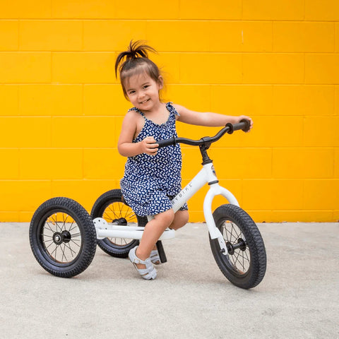 A young girl in a blue and white dress rides a white TryBike Steel tricycle from TryBike, featuring black tires and smiling at the camera. The background showcases a bright yellow wall, adding a vibrant pop of color to the scene.