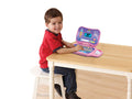 A young child wearing a red shirt and blue jeans sits on a white stool at a wooden table, smiling while using a Vtech Toddler Tech Laptop Pink with colorful buttons and display, perfect for developing early computer skills.