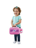 A young child with curly hair, wearing a light blue shirt and dark blue jeans, smiles while holding a pink Vtech Toddler Tech Laptop. The Vtech toy features a handle and a mesh-like front cover. The child stands against a white background, wearing white socks with no shoes.