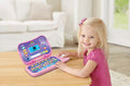 A young child with blonde hair and a pink shirt is sitting at a table, smiling at the camera. She is playing with a vibrant Vtech Toddler Tech Laptop Pink, which features a bright screen and various buttons. The cozy living room setting includes a white sofa and large windows in the background.