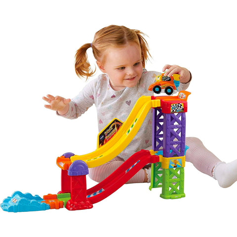 A young girl with pigtails plays with a vibrant plastic car ramp. The Vtech Toot Toot Drivers 3 in 1 Raceway, by Vtech, features a yellow slide, purple and green supports, and blue and red base elements. The child is holding a SmartPoint racer and looks engaged and happy.