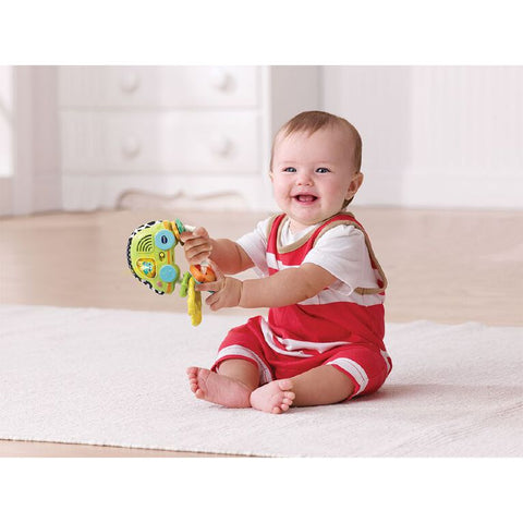 A baby wearing a red outfit sits on a white carpet in a brightly lit room, holding the colorful Vtech Touch & Feel Sensory Keys by Vtech with a smile. The background includes white drawers and a blurred wall, suggesting a cozy indoor environment.