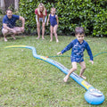 A group of four people, consisting of two adults and two children, delights in outdoor water play in a grassy yard. One child is running on a vibrant Wahu Splash 'N Snake by Wahu, with water jetting from the slide like a kids' sprinkler toy, while the others watch and smile.