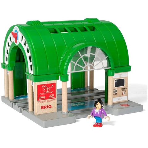 The Brio World Central Train Station set by Brio features a wooden toy train station with a green arched roof and gray tracks. Included in the set is a small, smiling figure of a person with black hair, wearing a purple top and blue pants. The central train station is adorned with various decals and logos, including one for "BRIO.