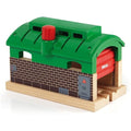 The Brio World Train Garage is a toy train engine shed made of wood and plastic, featuring a green roof with a red chimney, brick-patterned walls, and a red door adorned with the brand name "Brio." The base has two arches that allow toy trains to enter and exit.