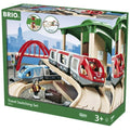 The image shows a boxed Brio World Travel Switching Set, suitable for ages 3 and up. The wooden set includes a battery-powered train, a bridge, tracks, and a figure. The box features the Brio logo and pictures of the assembled set with a cityscape backdrop.