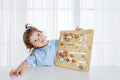 A young child with light brown hair in a small ponytail, wearing a light blue top, sits at a table holding up the Classic World 2 in 1 Pathfinder educational toy with various animal illustrations. The softly lit white curtains in the background add to the atmosphere of children's learning.