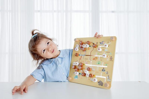 A young child with light brown hair in a small ponytail, wearing a light blue top, sits at a table holding up the Classic World 2 in 1 Pathfinder educational toy with various animal illustrations. The softly lit white curtains in the background add to the atmosphere of children's learning.