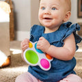 A happy baby with light skin and light hair is sitting on a carpeted floor, wearing a blue ruffled top and light-colored pants. The baby is holding the "Fat Brain Toys Dimpl" by Fat Brain Toys, a colorful sensory toy featuring four different silicone bubbles that promote tactile development.