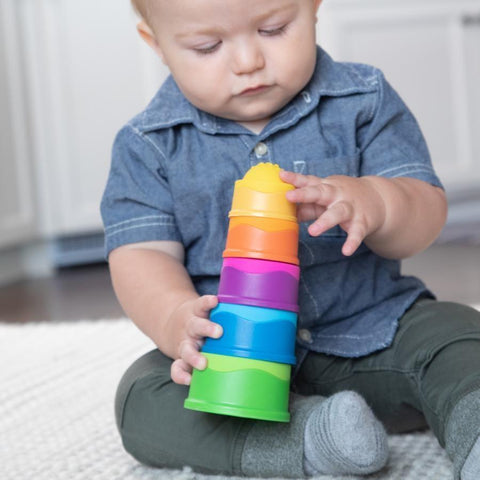 A toddler in a blue shirt and green pants sits on a white rug, engrossed in stacking colorful Fat Brain Toys Dimpl Stack cups, arranging them from largest to smallest. With tactile fascination, the child continues building while white kitchen cabinets are visible in the background.