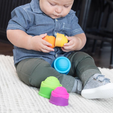 A baby wearing a blue shirt and green pants sits on a beige carpet, holding colorful Fat Brain Toys Dimpl Stack cups. The cups are green, purple, orange, and blue. Exhibiting tactile fascination with the toy, the baby is focused on stacking more Dimpl Stack cups from Fat Brain Toys around his feet.