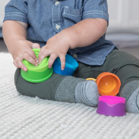 A child sits on a light-colored carpet, wearing a blue shirt and green pants, deeply engaged with their Fat Brain Toys Dimpl Stack. The child's face is not visible as green, blue, orange, and purple cups from the Fat Brain Toys collection are scattered around them.