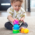 A young child with curly hair sits on a carpeted floor, intently stacking the vibrant cups from the Fat Brain Toys Dimpl Stack by Fat Brain Toys. The child is dressed in a white shirt adorned with a lizard pattern and dark pants. The background reveals blurred indoor elements, highlighting a home setting filled with tactile wonder.