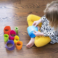 A child with blonde hair sits on a wooden floor, wearing a white shirt with black polka dots and yellow pants, deeply engaged in tactile discovery. She plays with Fat Brain Toys Spiroku from Fat Brain Toys, colorful stack-and-nest toy cups scattered around her, holding a blue cup in her hands and developing fine motor skills.