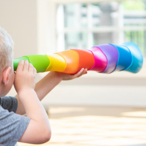 A child with blonde hair looks through a telescope made from the Fat Brain Toys Spiroku, a stack-and-nest toy of colorful, rainbow-hued plastic cups. The background is softly blurred, emphasizing the child's tactile discovery and creative play with the vibrant cups from Fat Brain Toys.