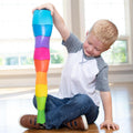 In a bright room with large windows flooding the space with natural light, a young boy with blonde hair and a gray and white shirt is seated on a wooden floor. He is engrossed in playing with his Fat Brain Toys Spiroku, carefully stacking the colorful, inverted cups into an impressive tall tower. The activity visibly delights him, enhancing his fine motor skills and encouraging tactile discovery.