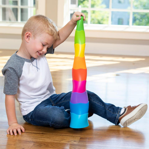 A young boy sits on the wooden floor of a brightly lit room, smiling as he engages in tactile discovery by stacking the colorful pieces of his Fat Brain Toys Spiroku into a tall tower. Wearing jeans, a white and gray raglan shirt, and sneakers, he hones his fine motor skills while sunlight streams through large windows in the background.