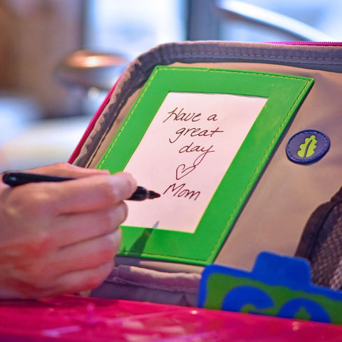 A hand holding a marker writes a note on a small white card framed in green, which reads "Have a great day ♥ Mom." The card is then tucked into the pocket of an insulated carry bag from the Go Green Value Bundle - Extreme by Go Green.