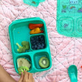 A child’s hand reaches for broccoli in a teal Goodbyn Hero (with Dipper Set) lunchbox, which is BPA free. The lunchbox contains mini bell peppers, blueberries, broccoli with dip, and a slice of kiwi. A teal spoon and compartment lid adorned with cartoon stickers rest nearby on a pink quilted blanket.