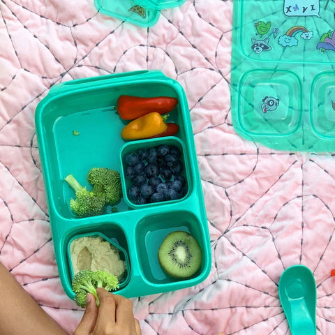 A child’s hand reaches for broccoli in a teal Goodbyn Hero (with Dipper Set) lunchbox, which is BPA free. The lunchbox contains mini bell peppers, blueberries, broccoli with dip, and a slice of kiwi. A teal spoon and compartment lid adorned with cartoon stickers rest nearby on a pink quilted blanket.