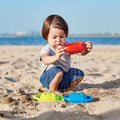 A young child with short brown hair is playing on a sandy beach, their little hands busy letting sand fall through an orange toy. In front of the child are HAPE Beach & Bath Boats, available in blue and green. The ocean and a clear sky create a beautiful backdrop for this moment of play, featuring products from HAPE.