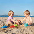 Two young children, a boy and a girl, are sitting on a sandy beach playing with HAPE Beach & Bath Boats from HAPE. The sea and a clear blue sky can be seen in the background. Both children are smiling and appear to be enjoying the sunny weather, their little hands busy with these colorful beach toys.