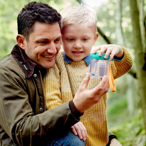 An adult and a child are outdoors, smiling as they examine the HAPE Explorers Bug Jar with its distinct green magnifying glass lid. The child, dressed in a yellow sweater, proudly holds up the HAPE bug jar to display something inside while the adult, wearing a brown jacket, looks on with pride. Trees can be seen in the background.