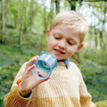 A young boy with blonde hair wearing a yellow sweater holds up the HAPE Explorers Bug Jar with a magnifying glass lid, examining it intently. The lush, green forest background suggests he is exploring nature.