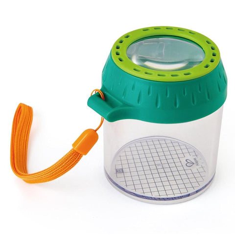 The HAPE Explorers Bug Jar by HAPE features a clear jar with a green-tinted magnifying glass lid, an orange carrying strap, and a gridded base inside for measurement. The lid has ventilation holes, while the bottom grid includes a small heart design. It is made from sustainable plastic.