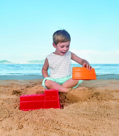 A young child sits on the sandy beach, immersed in imaginative role play with their HAPE Great Castle Walls mold. The ocean and blue sky provide a serene backdrop as the child enjoys the sunny day, creating adventures with their HAPE sand toys.