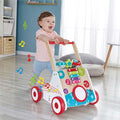 A toddler in a pink shirt and blue pants pushes the HAPE My First Musical Walker with adjustable speeds. This colorful walker from HAPE features musical notes, shapes, and a xylophone. The room has wooden flooring and minimalist decor bathed in natural light, creating the perfect setting for encouraging early walking skills.