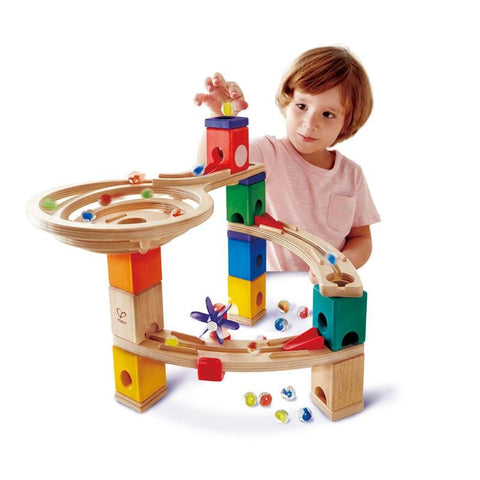 A young child with short brown hair, wearing a pink shirt, is playing with the HAPE Race to the Finish. This 58-piece beginner's set by HAPE features various colorful blocks and tracks, with marbles moving along the paths. The child is placing a marble at the top of the structure.