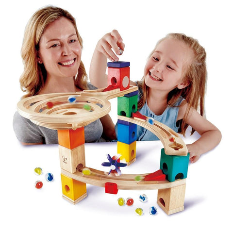 A smiling woman and a young girl are playing with the vibrant HAPE Race to the Finish set. The woman watches as the girl drops a marble onto the top track. Various marbles are scattered around the bottom of the 58-piece set, which features ramps, blocks, and spinners from HAPE.