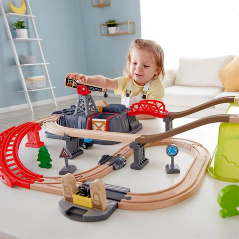 A young child with blonde hair sits at a table, playing with the HAPE Railway Bucket Builder Set. This 50-piece wooden train set includes tracks, bridges, and a crane, offering endless possibilities to build your own town. The background features a light blue wall and a white ladder shelf adorned with decorative items. The child is smiling and fully engaged.