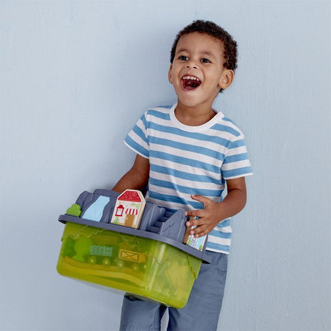 A young child with short curly hair smiles widely while holding a green HAPE Railway Bucket Builder Set, which contains various miniature toy buildings from the 50-piece collection. The child is wearing a blue and white striped t-shirt and gray pants, standing against a light blue background, ready to build their own town.