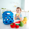 A young child sits at a table holding a toy banana, smiling. In front of the child are various soft fabric fruits, including an apple, a strawberry, a carrot, a pear, and a watermelon slice from the HAPE Toddler Fruit Basket by HAPE. A blue soft felt basket with circular holes rests on the left side of the table.