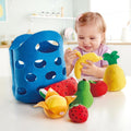 A toddler in a kitchen setting is playing with the HAPE Toddler Fruit Basket, which includes plush toy fruits like a banana, pineapple, strawberry, guava, and watermelon. Next to the child is a soft felt basket from HAPE with circular cutouts, holding the soft fabric fruits.