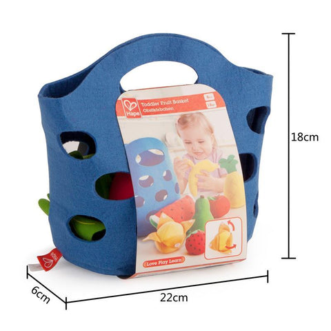 The HAPE Toddler Fruit Basket by HAPE includes a blue felt basket with handle holes, filled with soft fabric fruits in vibrant colors. The packaging features a photo of a child playing with the baby fruit toys and partially covers the basket. The product dimensions are 22cm in width, 18cm in height, and 6cm in depth.