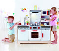 Two children engage in role-play with the Hape All-in-1 Kitchen by HAPE. The boy on the left, kneeling, is mixing something in a bowl while the girl on the right operates the toy microwave. The kitchen set, complete with accessories like a sink, stove, oven, and cabinets, is in a bright room with decorations.