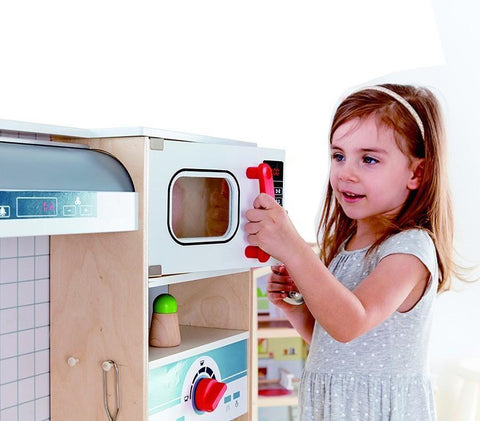 A young girl with light brown hair, dressed in a grey short-sleeve dress adorned with white polka dots, is role-playing at the HAPE All-in-1 Kitchen. She is holding the red handle of a white toy microwave door, smiling and peering inside, surrounded by various accessories.