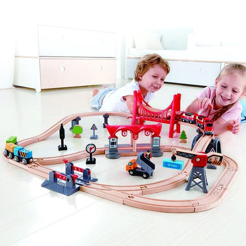 Two children lie on the floor playing with the Hape Busy City Rail Set, complete with Hape rail accessories. The city-themed rail set includes tracks, a red bridge, various signs, buildings, and toy vehicles. Both children are smiling and engaged in moving the passenger and freight trains around the tracks.