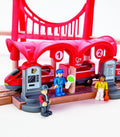 A city-themed rail set, known as the Hape Busy City Rail Set by Hape, features a red train at a station covered with a red canopy, marked by numbers 1 and 2. The set includes three toy figures: a conductor in a blue uniform, a worker wearing a red hat, and another worker dressed in yellow clothing. Additionally, the scene is adorned with red bridge arches and various rail accessories from Hape.