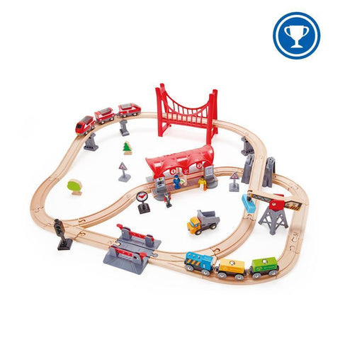 The Hape Busy City Rail Set by Hape is a wooden toy train set that features various tracks, bridges, and accessories. This city-themed rail set includes colorful passenger and freight train cars, a red suspension bridge, a gas station, a construction worker figure, a small tree, and various road signs. A blue trophy icon is displayed in the corner.