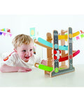 A young child with blonde hair and a white shirt is smiling while playing with the colorful Hape Fast Flip Racetrack, a delightful kids' toy from the brand Hape that features numbered tracks and small racing cars. The scene appears to be in a bright and cheerful room decorated with paper bunting.