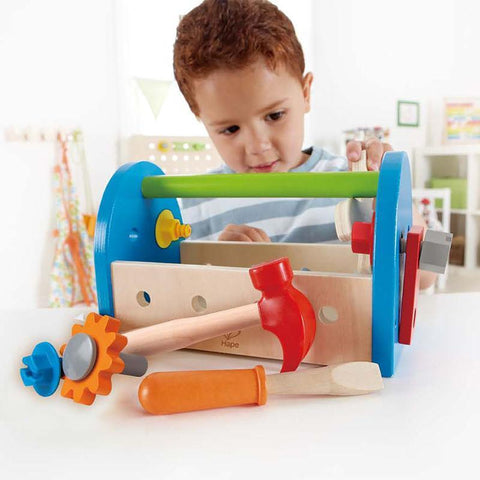 A young child is engrossed in the vibrant Hape Fix-It Tool Box from Hape, which includes a blue toolbox for kids and a variety of toy tools like a hammer, gear, screwdriver, and wrench. The child focuses on the toy at a white table, with a bright background highlighting various furniture.